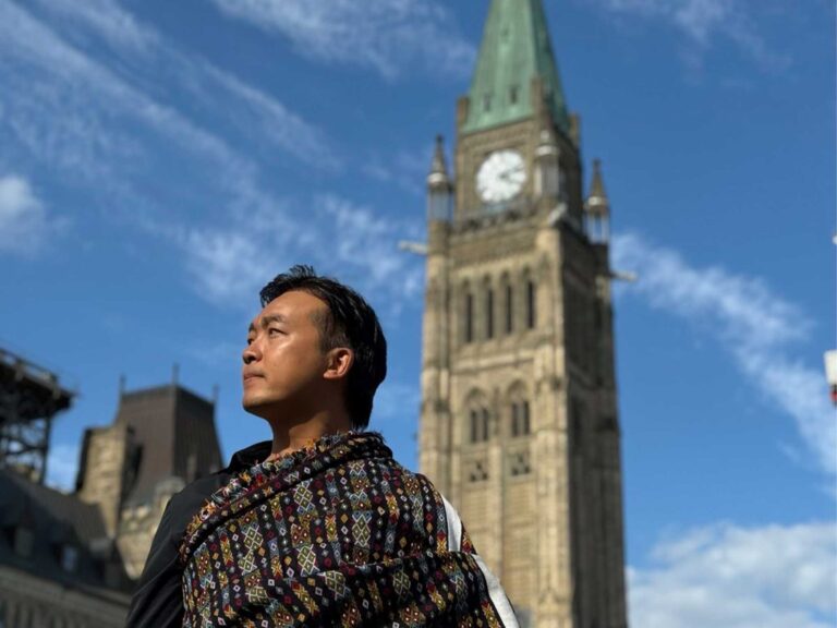 A person wearing a patterned woven garment stands outdoors with a tall clock tower in the background. The tower has a green pointed roof and ornate stone architecture. The sky is bright blue with scattered clouds.