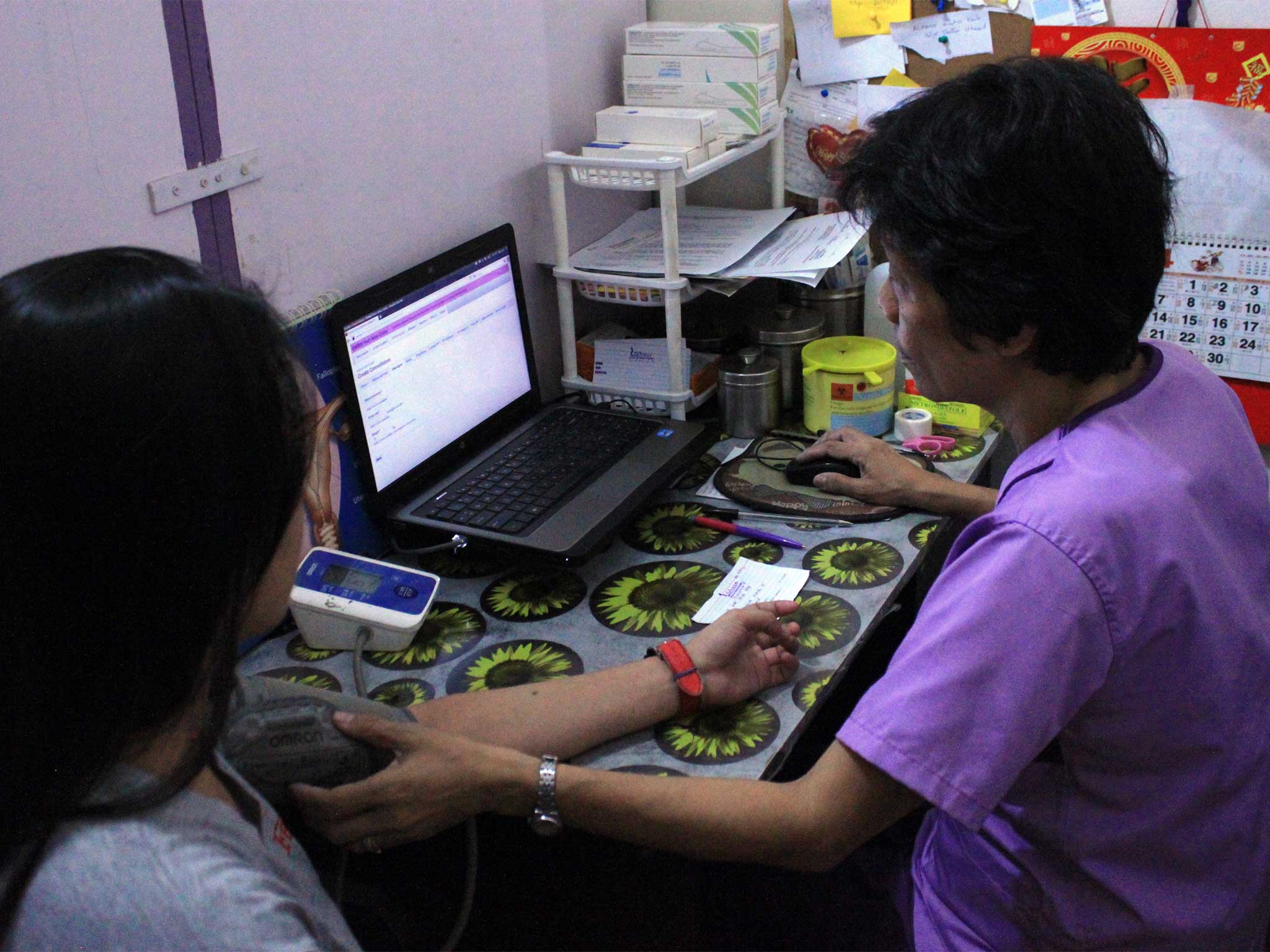 A healthcare worker seated at a small desk takes the blood pressure of a person sitting nearby. A laptop displaying medical information is open on the desk, which is covered with a sunflower-patterned tablecloth. Medical supplies, papers, and a calendar are visible in the background.
