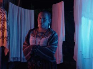 Person wearing patterned clothing stands between hanging textiles under dramatic blue and red lighting indoors, facing camera with folded arms.