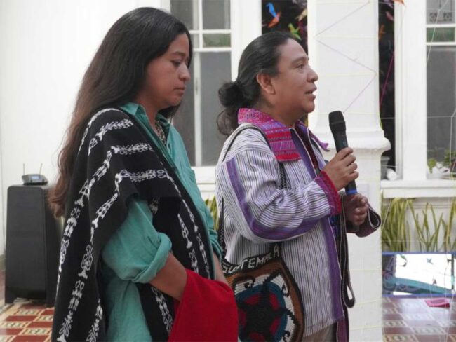 Two people stand, one speaking into a handheld microphone, wearing patterned garments during a community gathering event.